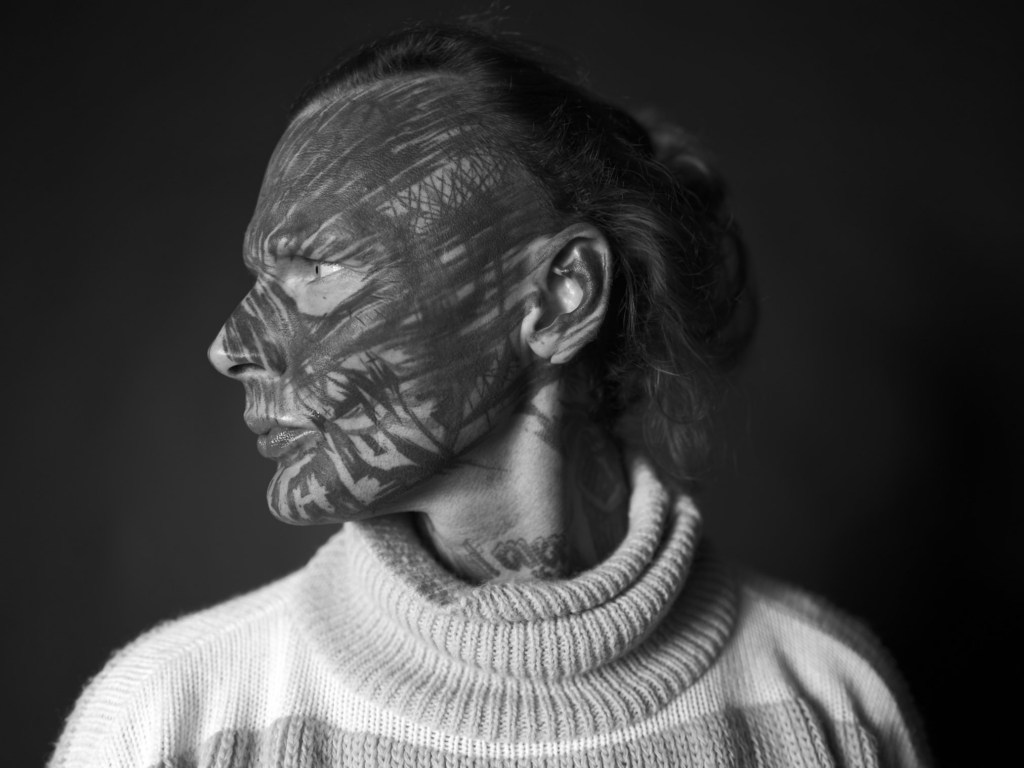 This is a black-and-white photograph by the photographer Konstantin Odin (model: Donnatella).
A side-profile portrait of a person with their hair tied back, looking away from the camera, with patterned light and shadow cast across the face.
The textured shadows create a layered, contemplative mood, highlighting facial contours against a dark, minimal background.