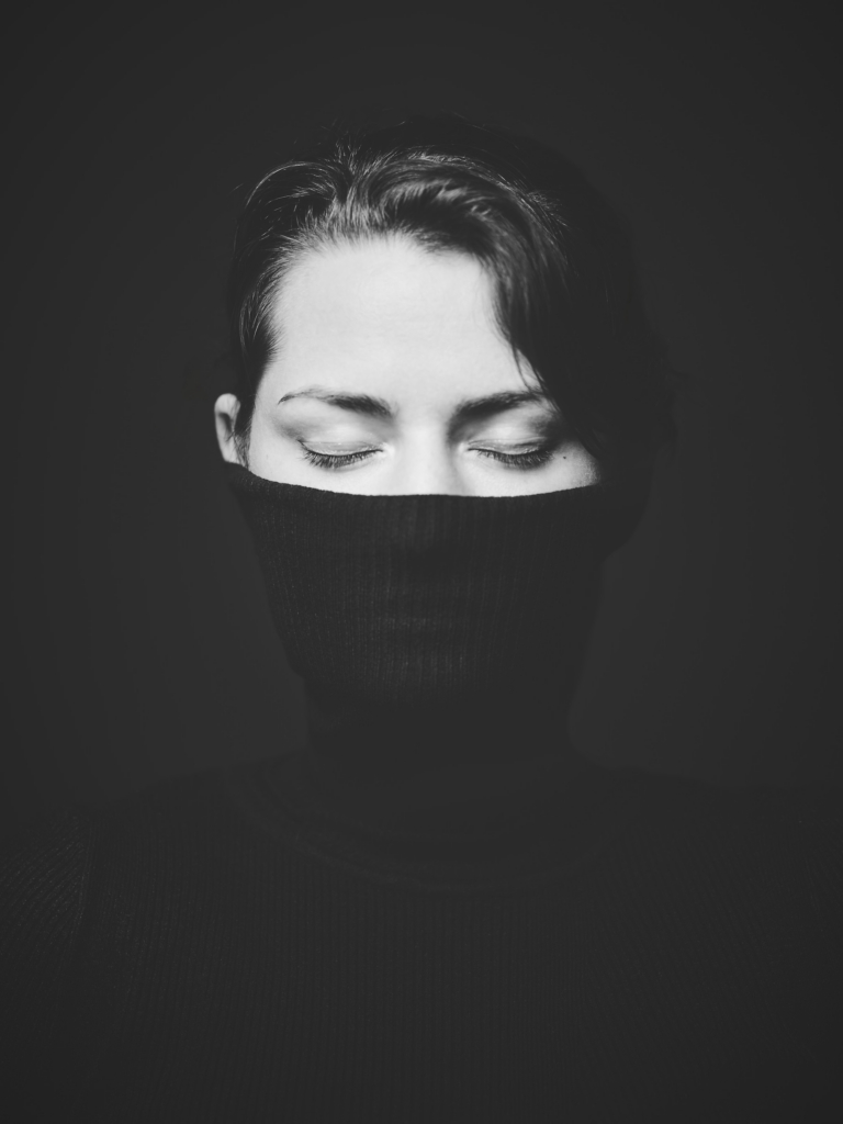 A black-and-white photograph by the photographer Konstantin Odin (model: Dorothea Adam).
A close-up portrait of a woman with her eyes closed, the lower half of her face covered by dark fabric.
Soft, dramatic lighting isolates her face against a deep black background, creating a quiet, introspective mood.