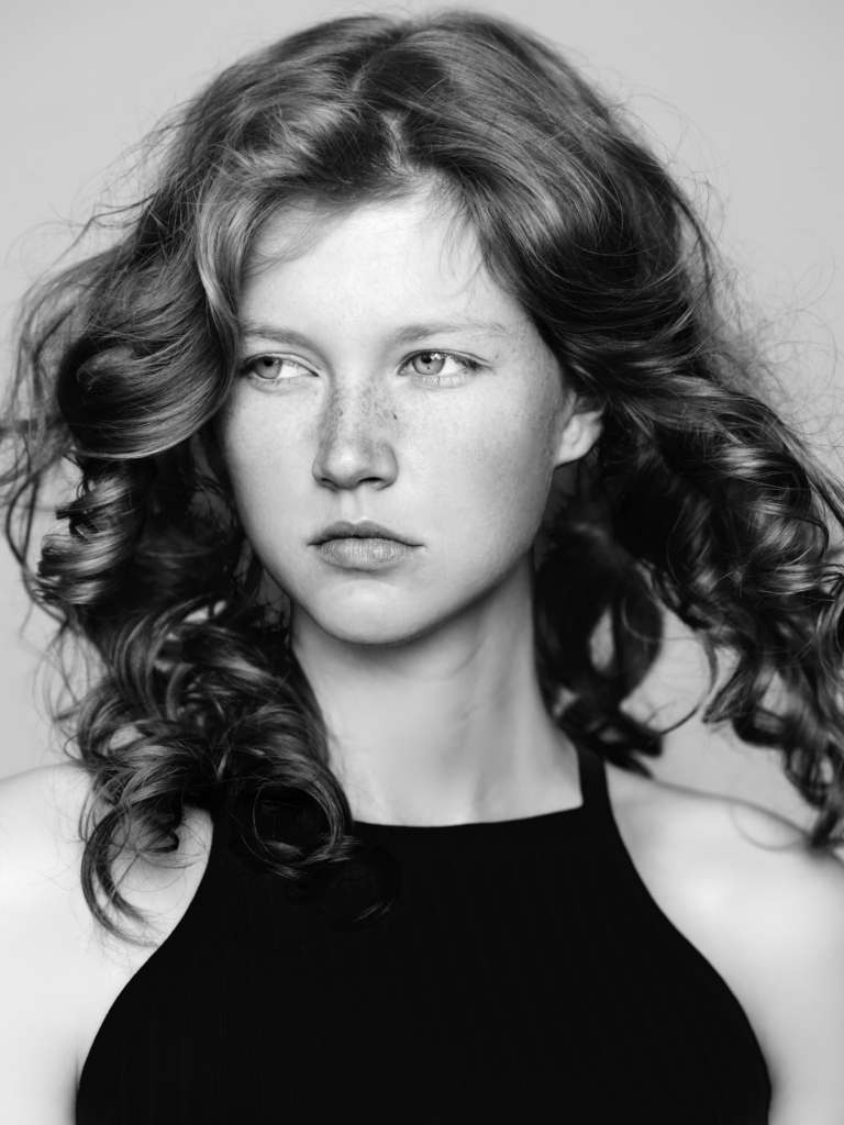 This is a black-and-white photograph by the photographer Konstantin Odin (model: Ellen Paul).
A young person with soft freckles and long, flowing hair looks slightly to the side, not directly at the camera.
The portrait is intimate and calm, with gentle light emphasizing facial features and texture of hair.