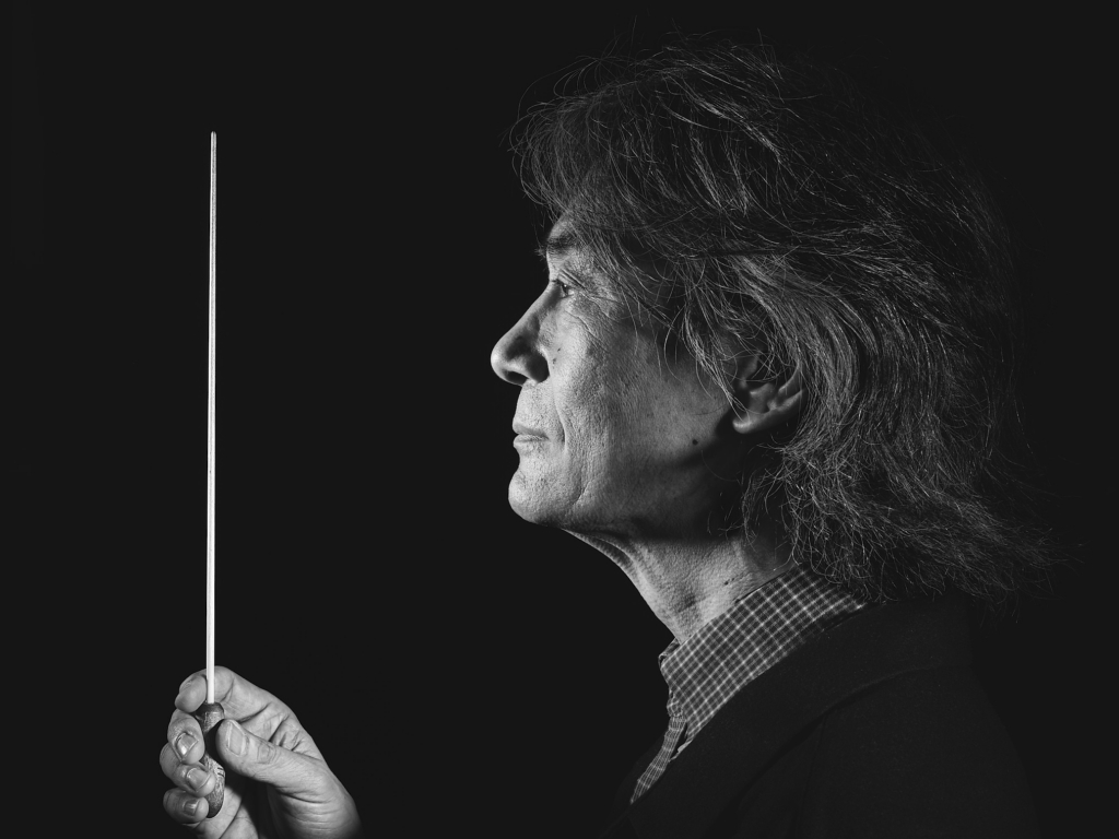 This is a black-and-white photograph by the photographer Konstantin Odin (conductor: Kent Nagano).
A side-profile portrait of a man holding a conductor’s baton upright in front of him.
The dramatic lighting highlights the texture of his hair and face against a deep black background, creating a focused, contemplative mood.