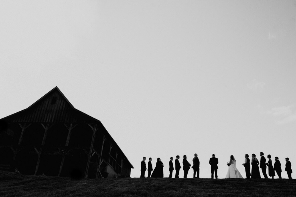 A line of wedding guests stands in silhouette on a hill, facing each other with the bride and groom near the center. The scene is photographed in black and white, with a large barn-like structure on the left and an open sky filling most of the frame. The wide composition emphasizes negative space and the graphic contrast between the dark figures and bright sky.