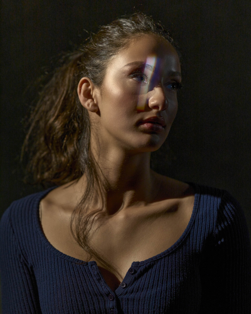 This is a color photograph by the photographer Konstantin Odin (model: Mira Hotlights).
A close-up portrait of a woman looking slightly to the side, with a soft beam of rainbow light crossing her face.
The dark background and gentle, directional lighting create a cinematic, intimate mood, drawing attention to her expression and the subtle play of color on her skin.