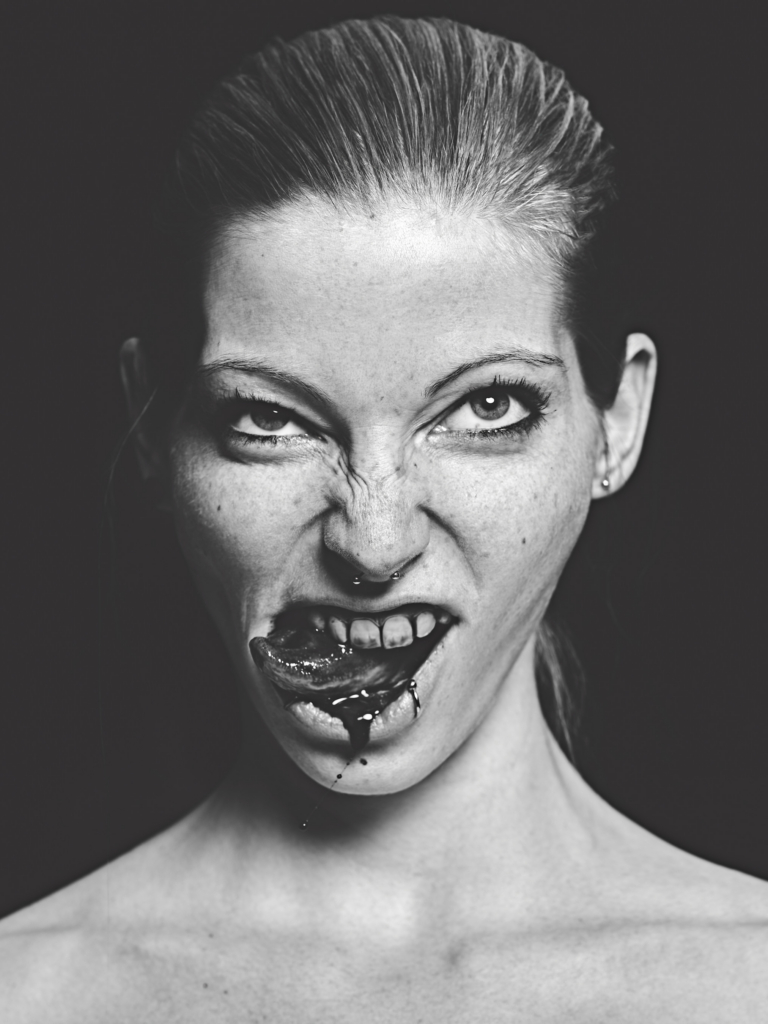 This is a black-and-white photograph by the photographer Konstantin Odin (model: Mirjam).
A close-up portrait of a woman making a fierce, expressive face, with dark liquid on her lips and chin.
Strong contrast and dramatic lighting emphasize texture and intensity against a deep black background, creating a raw, powerful mood.