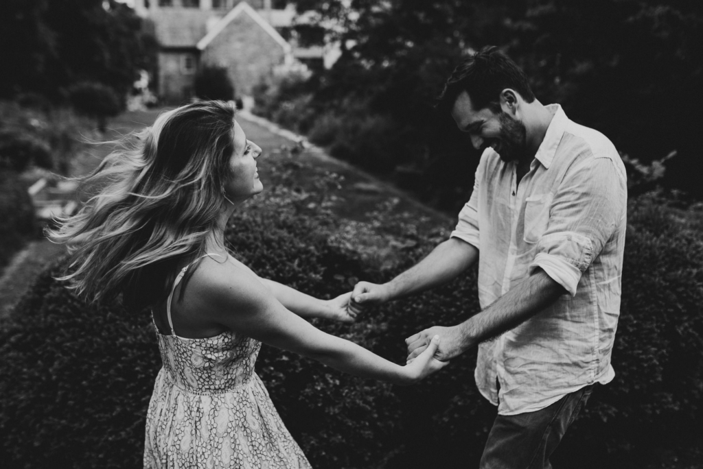 A couple holds hands and gently spins together outdoors, smiling and facing each other. The image is in black and white, with the woman’s hair and dress moving as she turns. A garden path and a house appear softly blurred in the background, giving the scene a candid, intimate feel.