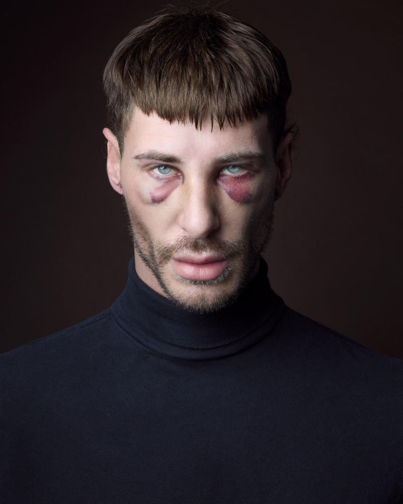This is a color photograph by the photographer Konstantin Odin (model: Pitzi Müller).
A close-up portrait of a person facing the camera, with visible marks beneath both eyes and an intense, direct gaze.
The dark background and soft, controlled lighting create a dramatic, intimate atmosphere that draws attention to the eyes and facial features.