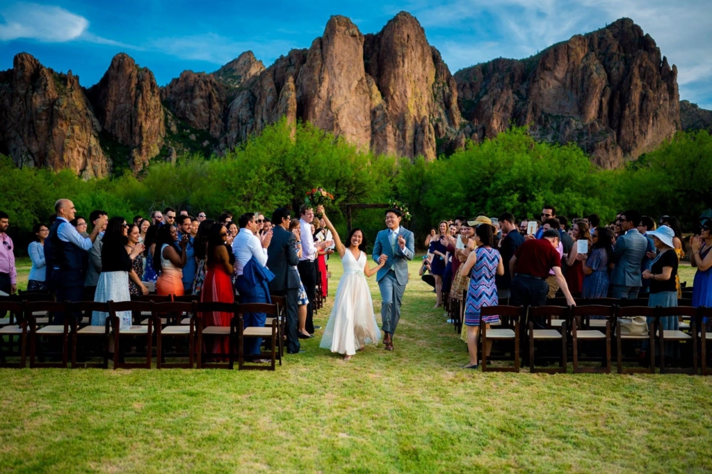 A newly married couple walks hand in hand down the aisle outdoors, smiling and raising their arms in celebration. Guests stand on both sides, clapping and taking photos as the couple passes. Behind them, dramatic rocky mountains rise above green trees under a bright blue sky.