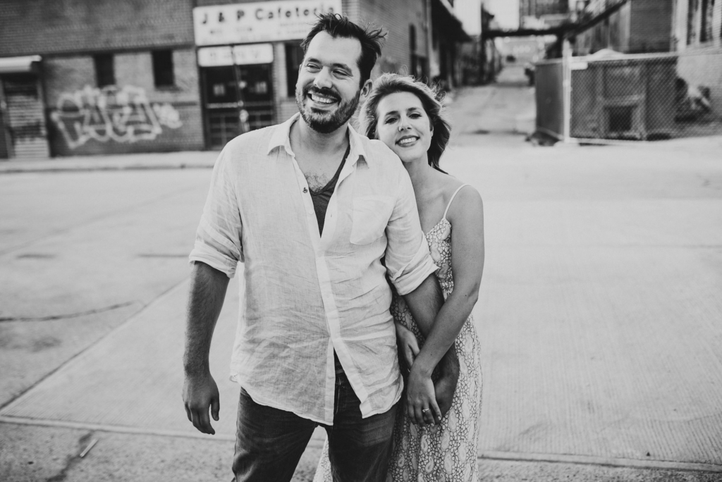 A couple walks together on a city street, smiling and relaxed, captured in black and white. The woman stands slightly behind the man, leaning into him with a warm expression. Urban buildings and graffiti appear softly in the background, giving the scene a casual, candid feel.