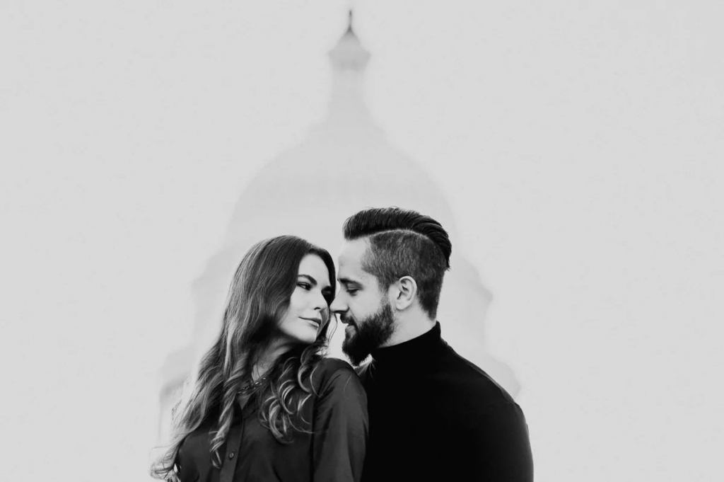 A couple stands close together, facing each other in a quiet, intimate pose. The image is in black and white, with a softly blurred domed building in the background. The shallow focus and minimal background draw attention to their faces and the closeness between them.