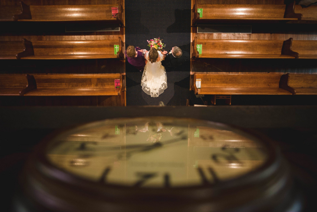 A top-down view inside a church shows a bride in a white dress walking down the aisle, holding a bouquet, with two people at her sides. Wooden pews line both sides of the aisle, creating a strong symmetrical pattern. In the foreground, a blurred circular element frames the scene, adding depth to the composition.