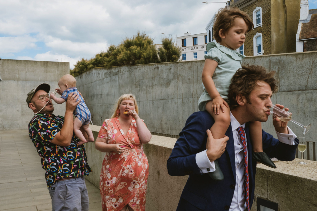 A group of wedding guests stand on a terrace: a man in a suit drinks champagne while carrying a child on his shoulders, another man lifts a baby in the air, and a woman in a floral dress eats a snack nearby.