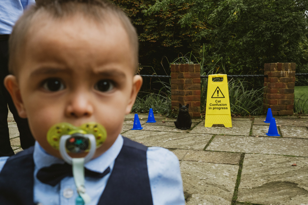 A toddler in a bow tie and vest looks into the camera with a pacifier, while a black cat sits behind him near a yellow warning sign reading “Cat confusion in progress” and small blue cones on a stone path.