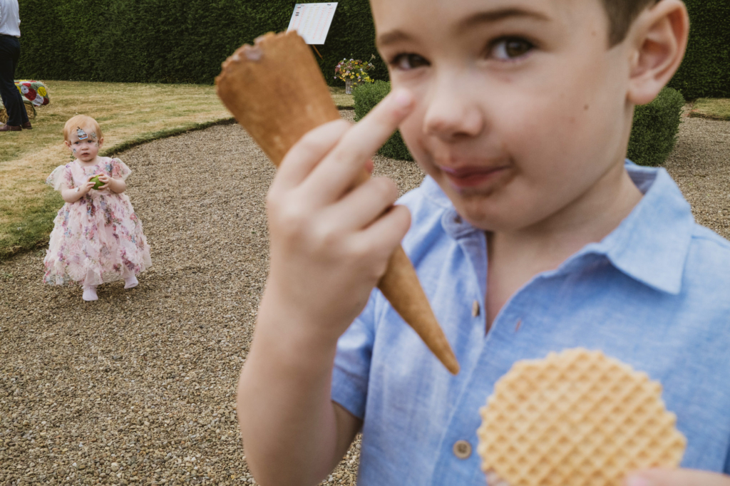 A young boy in a blue shirt holds an ice cream cone close to his face while a toddler in a floral dress stands behind him on a gravel path in a garden.
