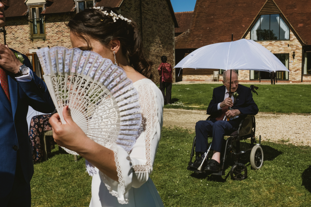 A bride holds a decorative hand fan in the foreground while an elderly man in a wheelchair sits under a white umbrella on the lawn outside a brick wedding venue.