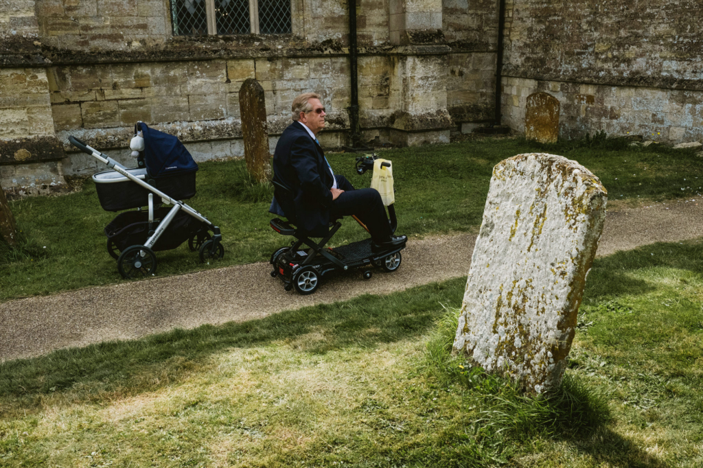 A man in a suit riding a mobility scooter along a church path, passing a baby stroller and gravestones in a quiet churchyard.
