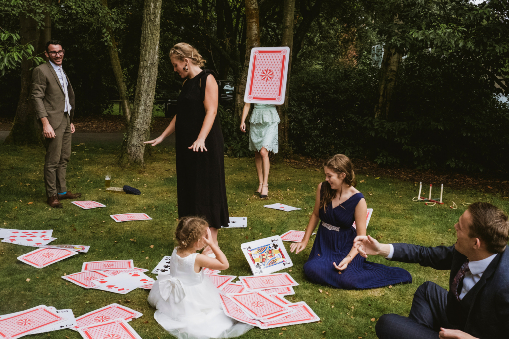 Wedding guests and children gather on a grassy lawn where oversized playing cards are scattered across the ground.
A young flower girl in a white dress sits among the cards, while adults in formal outfits laugh and gesture around her.
The scene feels playful and spontaneous, like a game unfolding in a garden during the celebration.