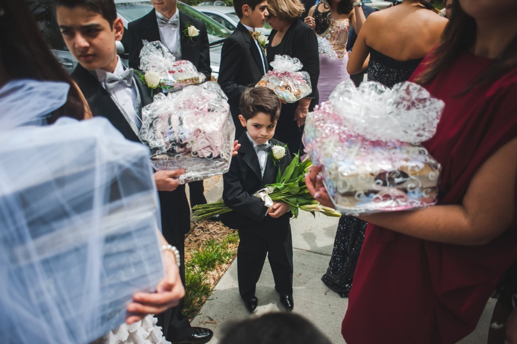 A young boy in a formal suit stands among wedding guests, holding a bouquet of white flowers. People around him carry wrapped gift baskets and wear formal attire, creating a busy, crowded scene. The moment feels candid and slightly chaotic, capturing the child’s small presence within the larger gathering.