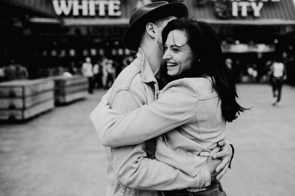 A couple embraces closely in a busy outdoor urban setting, captured in black and white. The woman smiles broadly while the man holds her, his face partly turned away. Blurred figures and storefronts in the background suggest a lively public space, adding a candid, spontaneous feel.