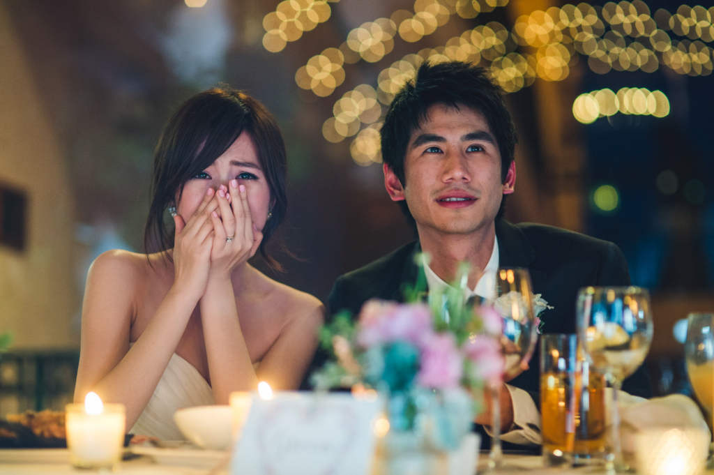 A bride and groom sit side by side at a reception table, lit by warm candlelight. The bride covers her mouth with both hands, appearing emotional, while the groom looks ahead with a soft smile. Out-of-focus string lights glow in the background, creating a warm, intimate atmosphere.