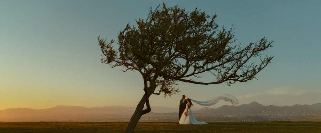 A bride and groom stand on a hill beneath a large windswept tree overlooking the city of Santiago, Chile. The bride’s veil flows dramatically in the wind while warm sunset light spreads across the mountains and skyline.

The wide cinematic composition emphasizes the scale of the landscape, placing the couple within the quiet beauty of the open horizon.