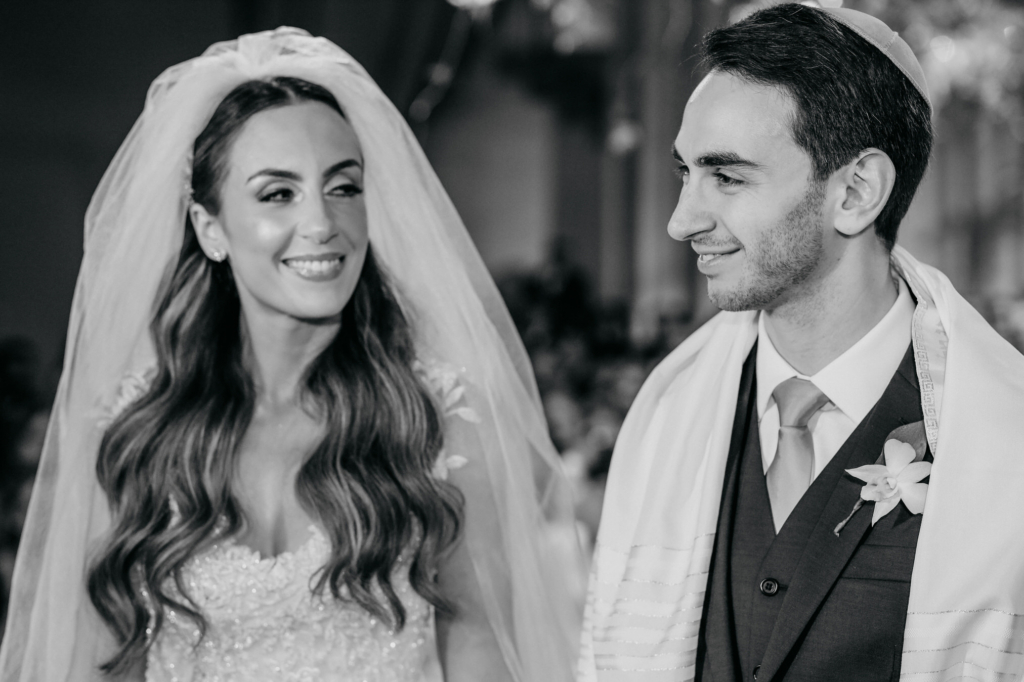 A bride and groom smile at each other during a wedding ceremony, both dressed in formal attire.