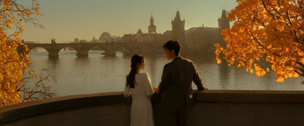 A couple stands side by side on a stone terrace overlooking the Vltava River in Prague. The historic Charles Bridge and old city towers appear in the distance, softly lit by warm autumn sunlight.

Golden leaves frame the scene as the couple looks at each other, creating a quiet, cinematic moment within the timeless landscape of the city.