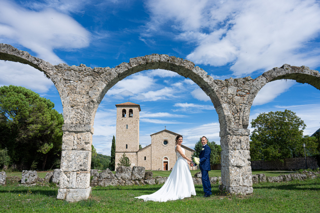 Bride and groom holding hands beneath ancient stone arches with a church in the background.