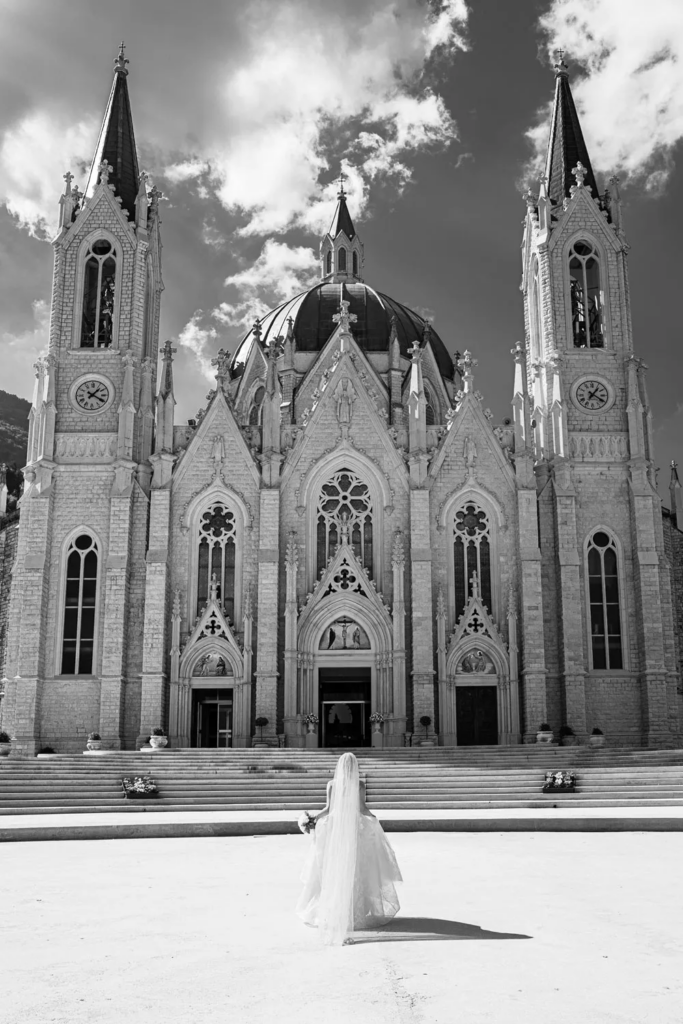Bride standing alone before a grand cathedral, captured in a dramatic black-and-white composition.