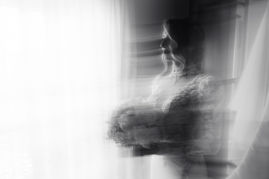 Blurred bride standing by a window, soft light and motion creating a dreamy, emotional atmosphere.