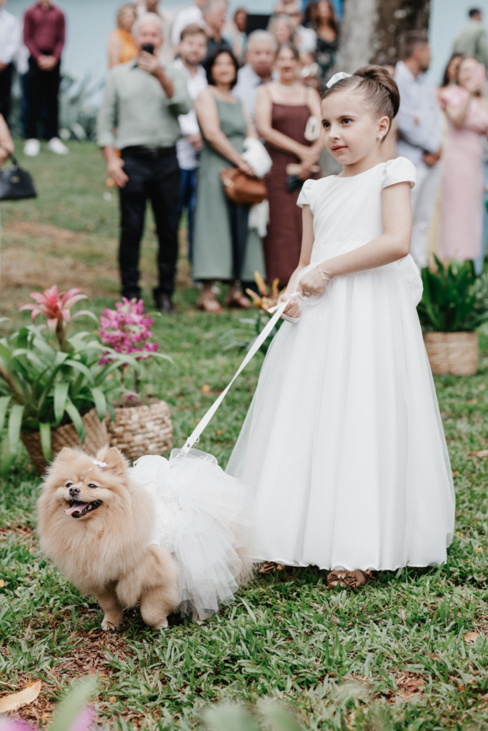 A young girl in a white dress holds a small dog on a leash during an outdoor wedding, with guests blurred in the background.