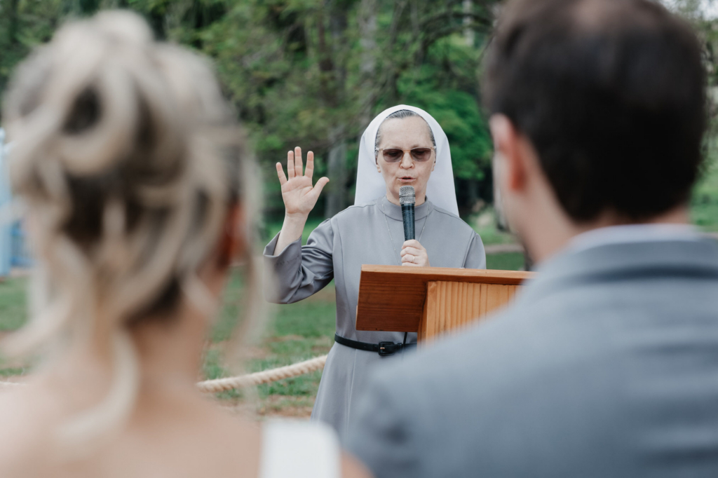 A nun stands outdoors holding a microphone and raising her hand while officiating a wedding ceremony, with a couple blurred in the foreground.