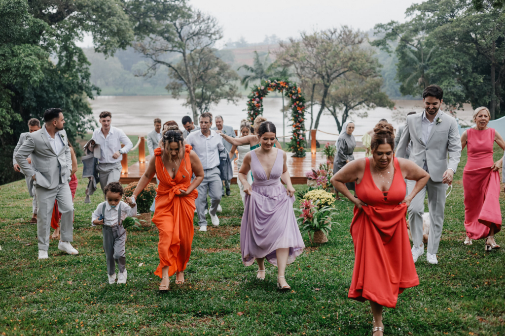 A group of wedding guests runs together across a grassy area outdoors, with a decorated ceremony arch and a lake visible in the background.