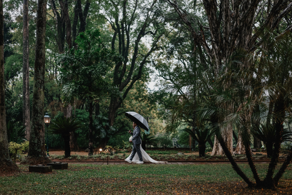 A couple walks together under an umbrella through a lush garden, surrounded by tall trees and greenery.