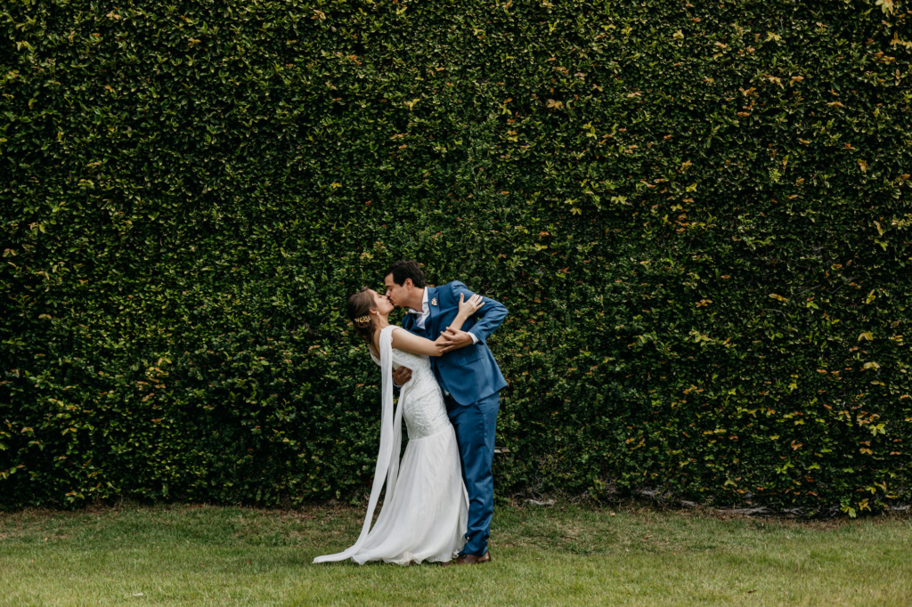 A couple in wedding attire shares intimate and joyful moments together outdoors, standing on grass in front of a lush green wall.