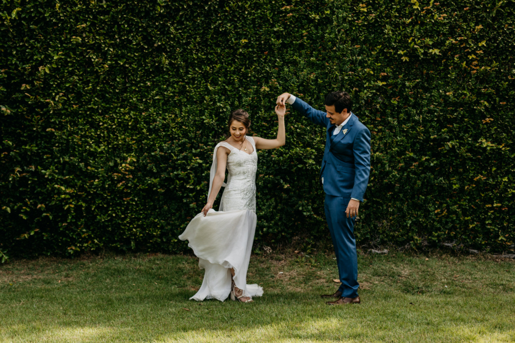 A couple in wedding attire shares intimate and joyful moments together outdoors, standing on grass in front of a lush green wall.