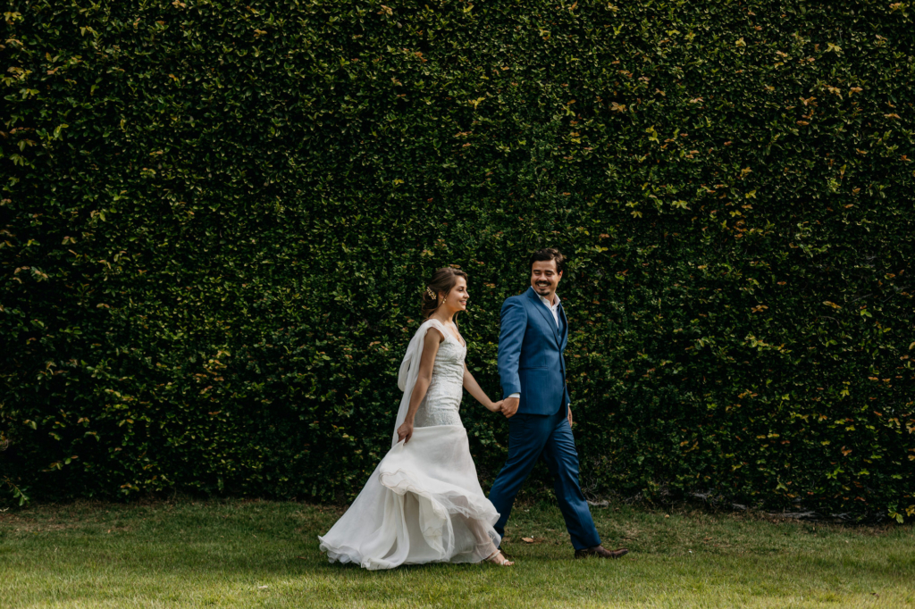 A couple in wedding attire shares intimate and joyful moments together outdoors, standing on grass in front of a lush green wall.