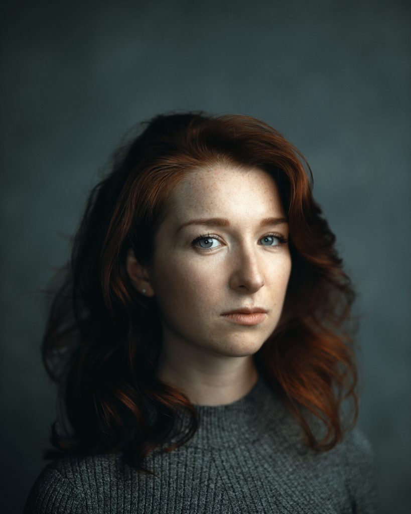 A close-up studio portrait of a woman with shoulder-length red hair and fair, freckled skin. She wears a simple gray knit top, and her blue eyes look directly into the camera with a calm, steady expression. Soft, directional light from one side gently shapes her face, creating a quiet, intimate atmosphere against a muted blue background.