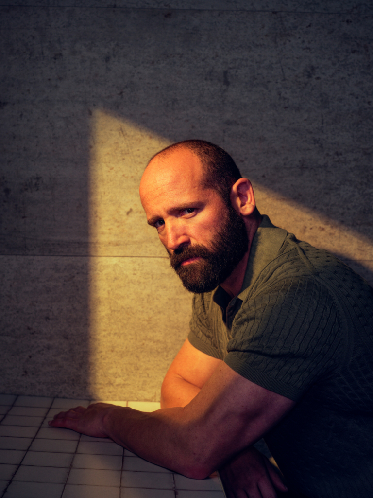A studio portrait of a bald man with a full beard leaning on a tiled surface, his forearms resting in front of him. A strong beam of warm light cuts across the wall and partially illuminates his face, creating dramatic contrast with the surrounding shadows. He looks toward the camera with a serious, contemplative expression, the textured wall behind him adding to the moody, cinematic atmosphere.
