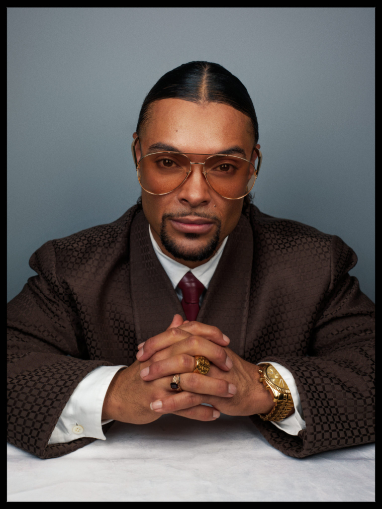 A studio portrait of a man seated at a table, his hands clasped together in front of him. He wears a patterned brown suit, a white shirt, and a deep red tie, along with large gold-tinted aviator glasses. Gold rings and a watch catch the light, while he looks directly into the camera with a composed, confident expression against a soft gray background.