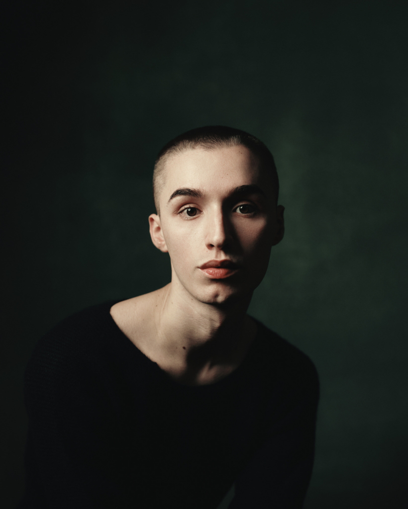 A studio portrait of a young person with a closely shaved head, wearing a simple black top. They face the camera directly, their expression calm and open, with soft light falling across one side of their face and fading gently into shadow on the other. The deep green background and controlled lighting create a quiet, intimate mood that draws attention to their eyes and subtle features.
