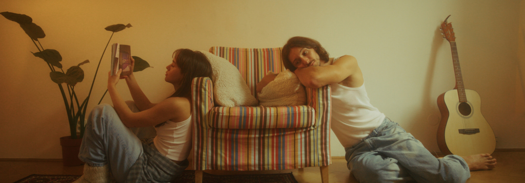 A young couple sits on opposite sides of a striped armchair in a softly lit room. The woman reads a book beside a tall houseplant while the man leans on the chair, resting his head on his arms and gazing toward her.

Warm golden light fills the quiet interior scene, with a guitar resting against the wall, suggesting an intimate and reflective moment between them.