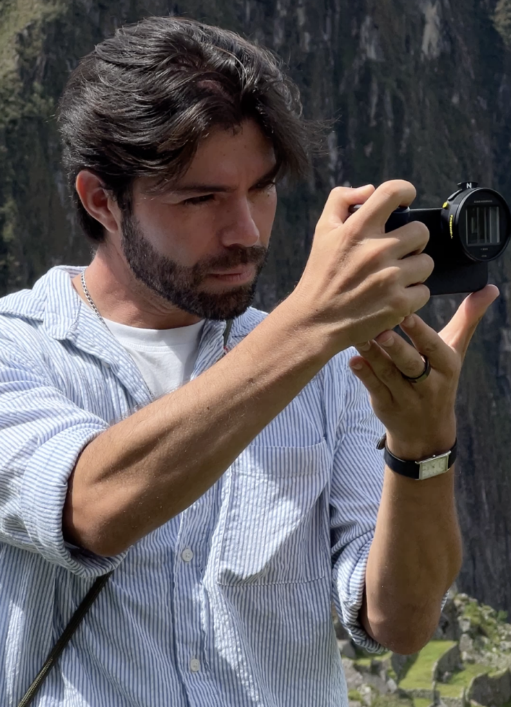 Portrait of photographer Braulio Lara concentrating as he holds a camera and frames a shot. He stands outdoors in bright daylight, focused on capturing an image.

Behind him, steep green mountains and rocky cliffs create a dramatic natural backdrop, reflecting his connection to landscape and cinematic composition.