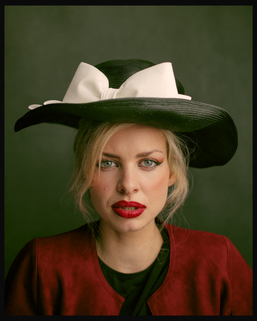 A close-up studio portrait of a woman facing the camera directly. She wears a wide-brimmed black hat adorned with a large white bow, a deep red jacket, and bold red lipstick that contrasts with her pale skin and light blonde hair. Her green eyes are sharply focused on the viewer, and the softly lit green background enhances the dramatic, painterly quality of the image.