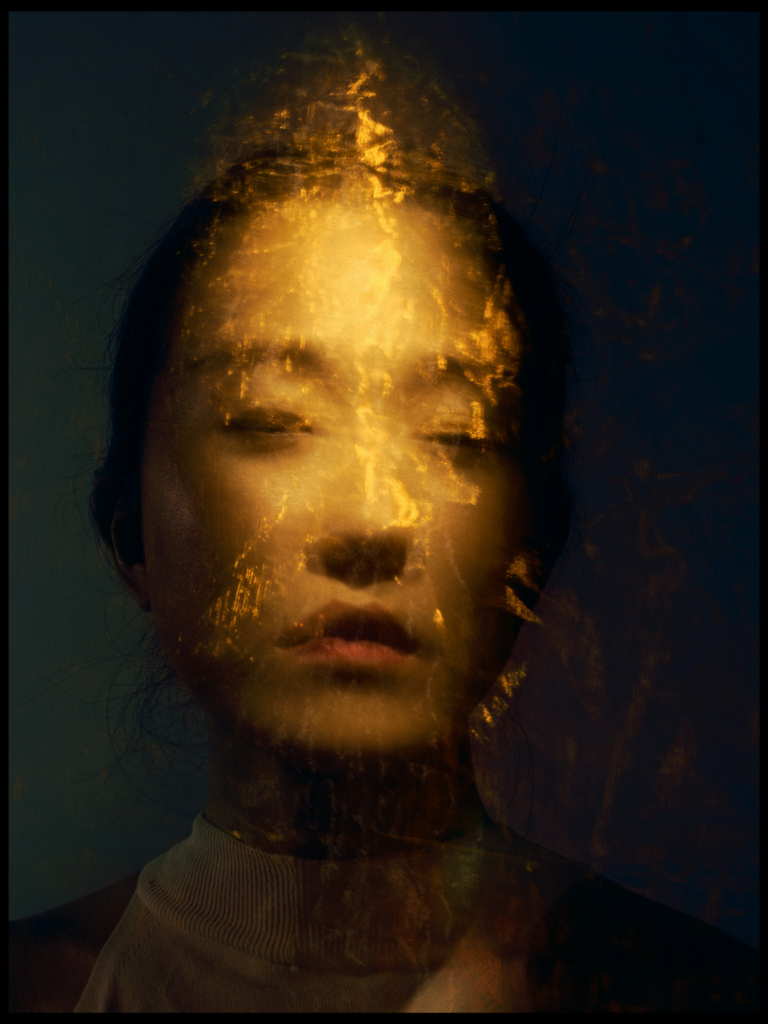 A close-up studio portrait of a woman with her eyes gently closed, her face partially obscured by a glowing overlay of golden light and textured reflections. The warm streaks of light flow vertically across her features, contrasting with the dark, cool-toned background. The layered effect creates a dreamlike, abstract atmosphere, blending her calm expression with luminous, almost flame-like patterns.