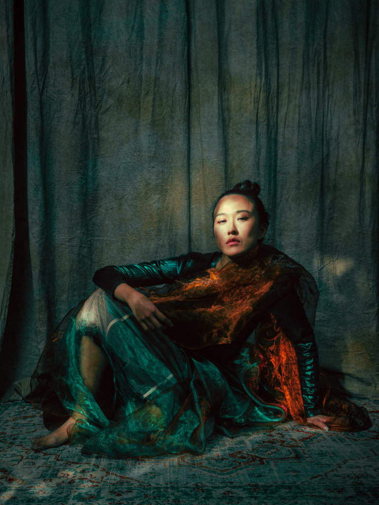 A dramatic studio portrait of a woman seated on the floor in front of heavy, textured curtains. She wears a dark, glossy top and is wrapped in flowing layers of translucent green and copper-toned fabric that catch the light like embers against deep shadows. Her posture is relaxed yet powerful, one arm supporting her as she gazes toward the camera with a calm, commanding expression, while moody lighting creates a cinematic, almost theatrical atmosphere.