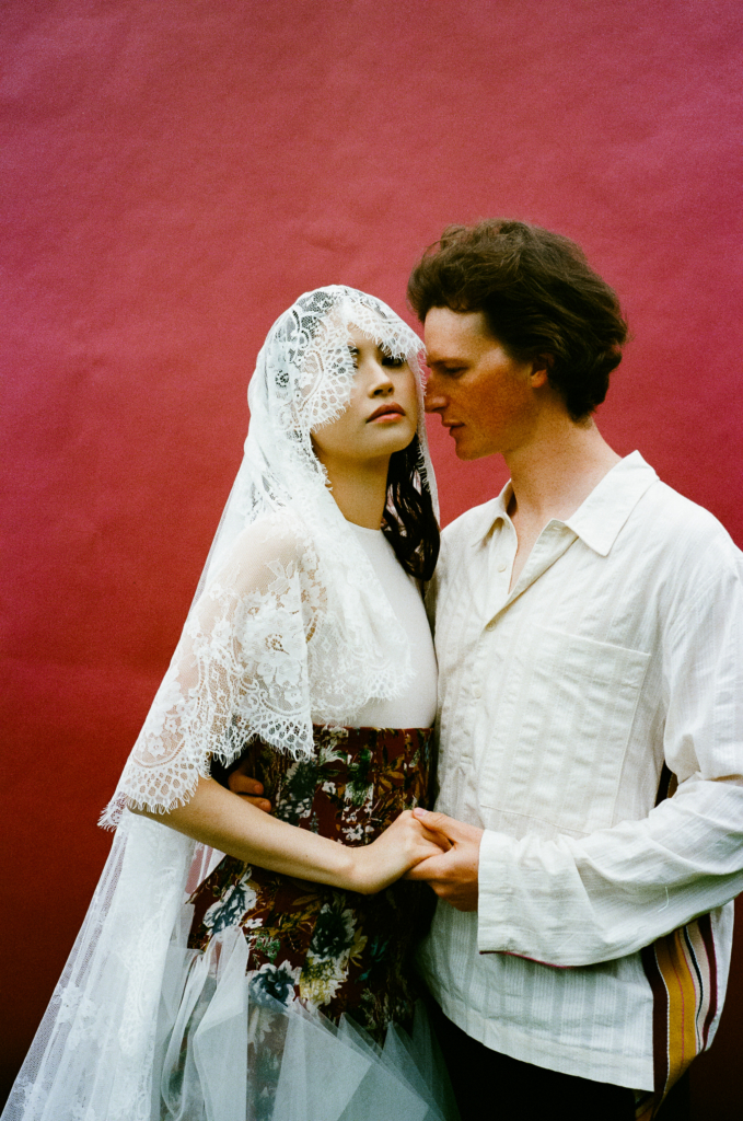 A woman in a lace veil and floral dress stands close to a man in a white shirt, holding hands against a deep red background.