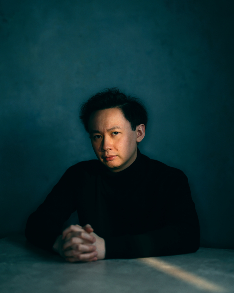 A studio portrait of a man seated at a table, his hands gently clasped in front of him. He wears a black high-neck sweater, and his face is softly illuminated from one side, creating subtle shadows that add depth and mood. The cool, muted blue background and controlled lighting give the image a quiet, contemplative atmosphere.