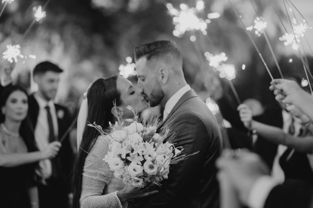 A couple kisses while holding a bouquet, surrounded by people holding sparklers in a softly lit nighttime scene.