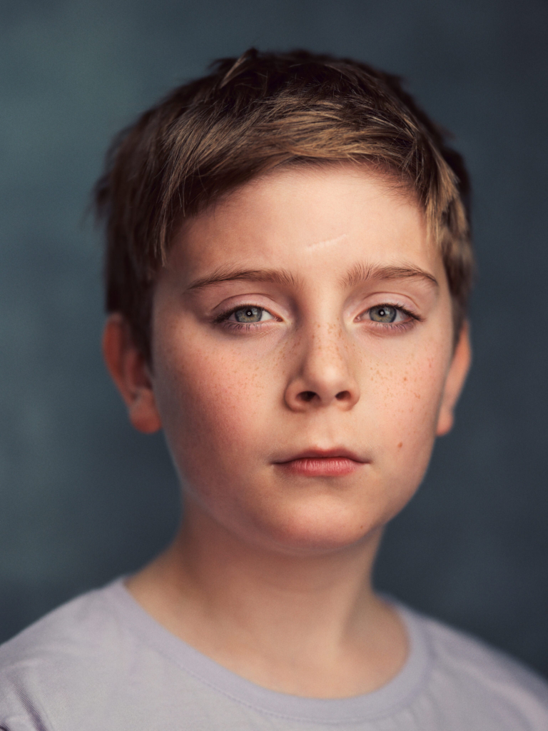 A close-up studio portrait of a young person with short light-brown hair and fair, freckled skin. Their blue eyes look directly into the camera with a calm, steady expression, lips relaxed and face softly illuminated from the side. The muted blue background and gentle lighting create a quiet, intimate mood that highlights the natural texture of their skin and subtle features.