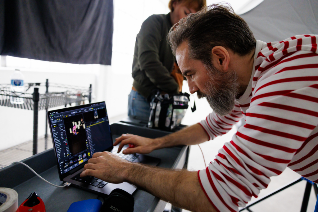 A behind-the-scenes image of Ivan Weiss leaning over a laptop in his studio, carefully reviewing images on the screen. He wears a red-and-white striped shirt, his focus intense as one hand rests on the keyboard and the other navigates the trackpad. Studio equipment and a colleague appear in the background, capturing the working rhythm of a portrait session in progress.

Shot by Rich Soublet @richsoublet2