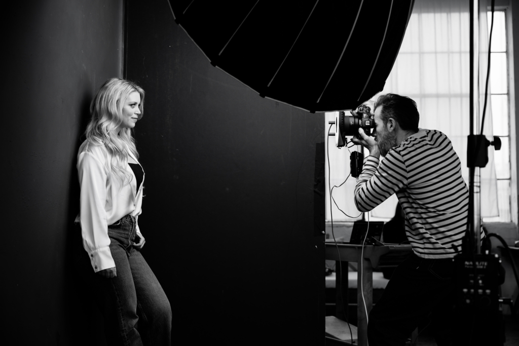 A black-and-white behind-the-scenes photograph of Ivan Weiss working in his studio, leaning forward as he photographs a woman standing against a dark wall. A large softbox light hangs above them, shaping the light across her face while he looks through the camera with focused concentration. The scene captures the quiet intensity of a portrait session in progress.

Shot by Rich Soublet.